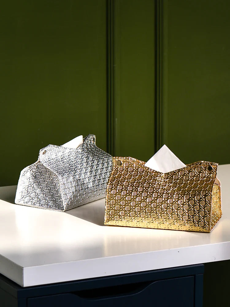 Two geometric tissue box covers, one silver and one gold, sit on a white countertop in front of a green wall. Each Glymmer Tissue Box holds tissues neatly protruding from the top, adding a modern decorative touch to your space.