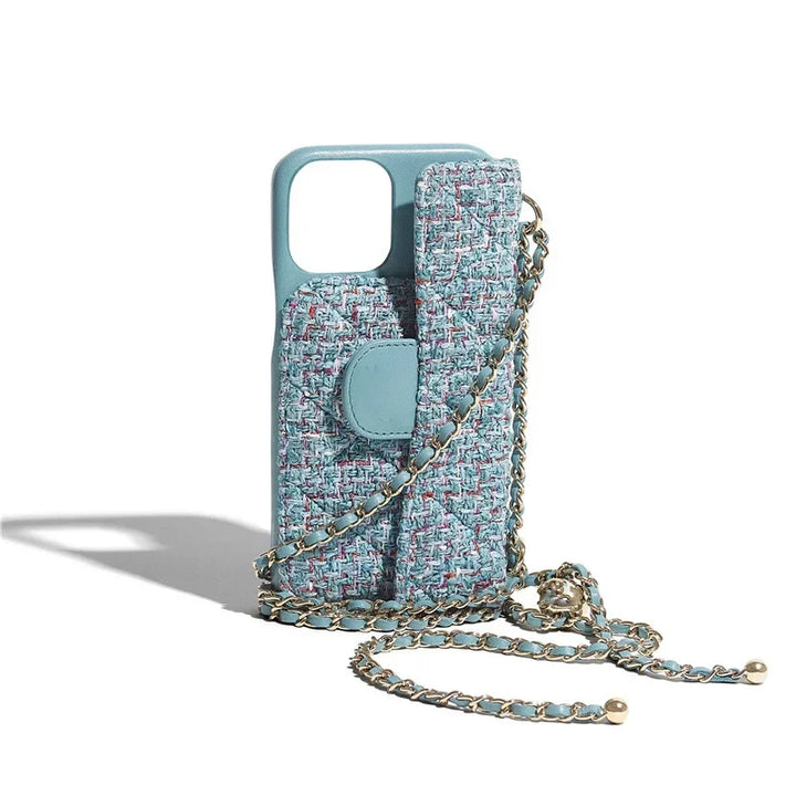 A stylish blue and pink tweed quilted phone case with a matching strap and gold chain accents, designed to be worn crossbody. The case is standing upright with the stylish chain strap laid out in front.