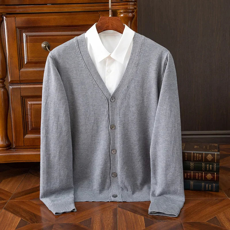 A light gray cashmere wool blend button-up cardigan with a relaxed fit is displayed on a hanger over a white collared shirt, set in front of a wooden dresser with books stacked nearby on a wooden floor.