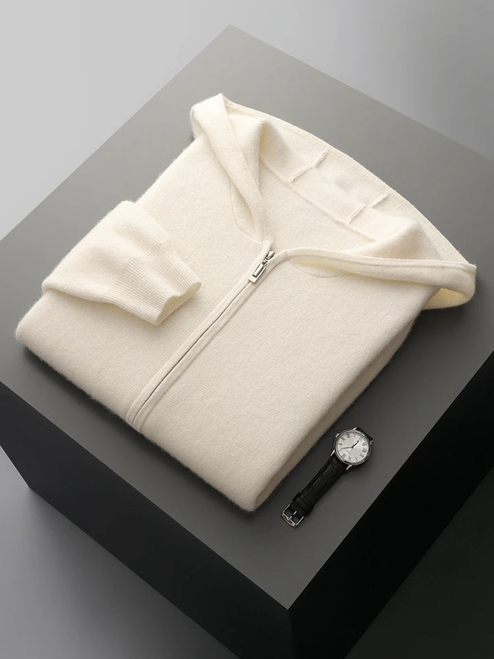 A cream-colored zip-up hoodie is neatly folded on a dark gray surface next to a black leather wristwatch with a white face, creating a cozy autumn winter cardigan vibe.