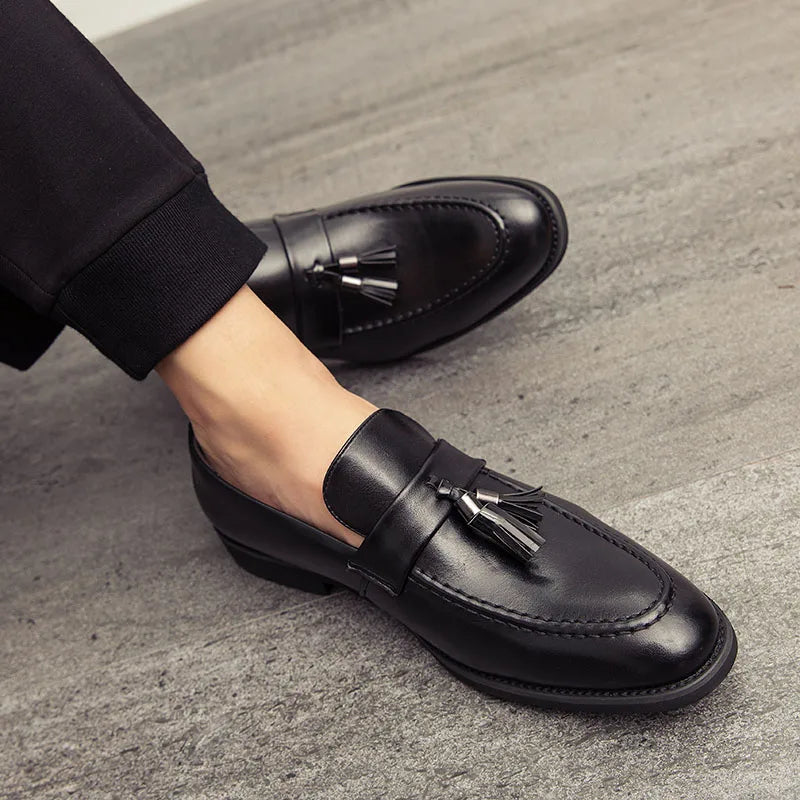 A person wearing black polished leather tassel loafers and black pants sits on a grey textured floor, with one leg extended and the other bent.