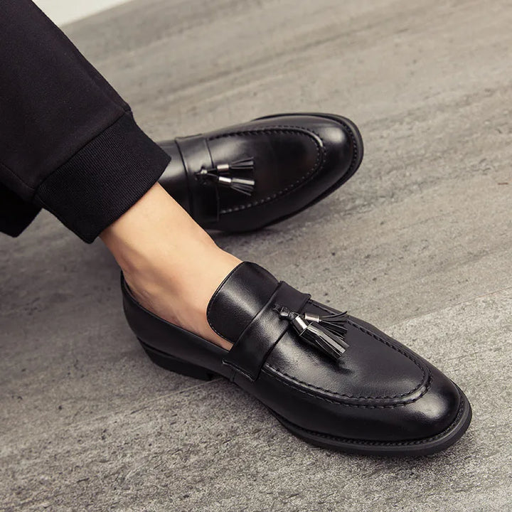A person wearing black polished leather tassel loafers and black pants sits on a grey textured floor, with one leg extended and the other bent.