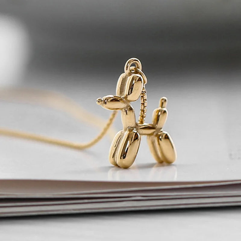 A gold balloon dog necklace with a pendant shaped like a balloon animal dog rests on a stack of white papers. The playful jewelry stands out against the softly blurred background.
