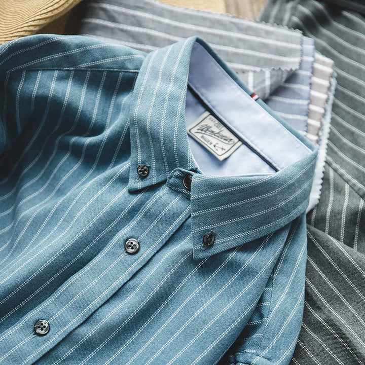 A close-up of several collared, button-up shirts with vertical stripes in blue and gray tones, crafted from fine weave cotton. Neatly layered, each pinstripe shirt features visible buttons and labels inside the collar.