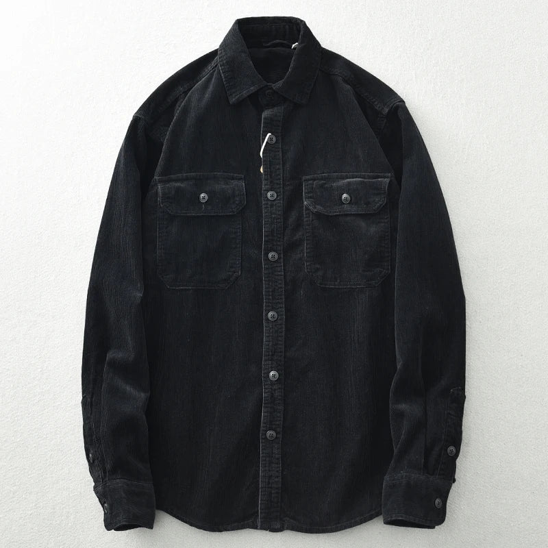 A black button-up shirt with a collar and two front chest pockets, crafted from heavyweight cotton and featuring corduroy detailing. Laid flat on a light-colored surface, the shirt has buttons down the front and on the cuffs.