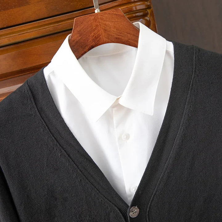 A white collared shirt is layered under a dark gray, relaxed fit V-neckline cardigan, both hanging on a wooden hanger in front of a wooden surface.