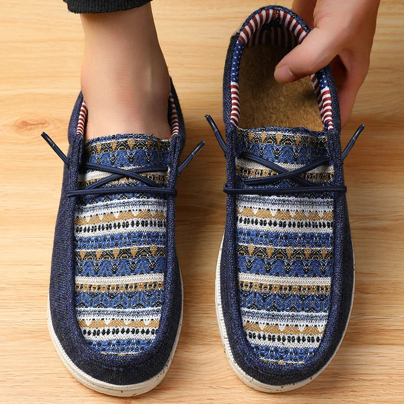 A person wearing blue slip-on shoes with a woven upper and vibrant ethnic pattern, featuring white soles, stands on a wooden floor. One hand adjusts the right shoe, which has geometric designs and a striped inner lining.