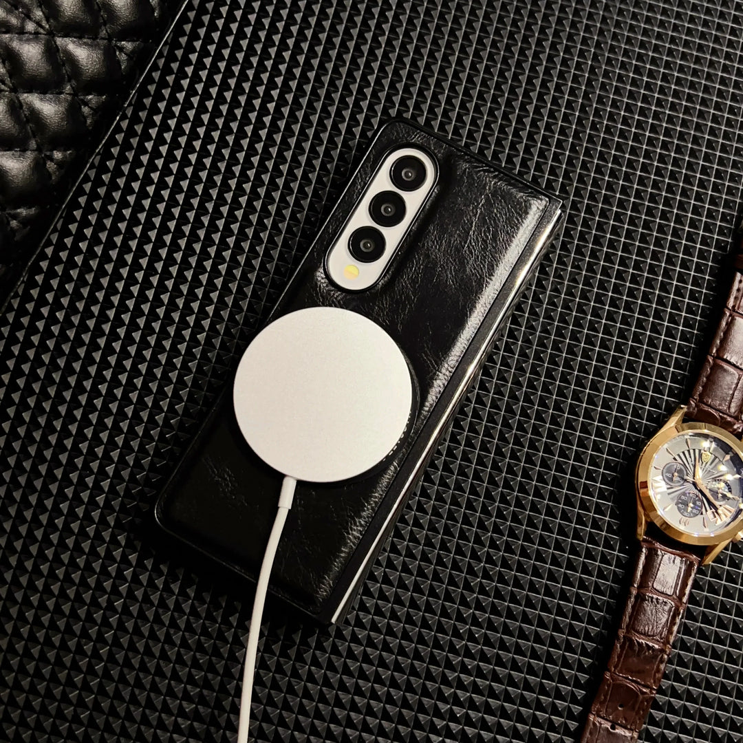 A smartphone with a black leather case is charging wirelessly via a round white MagSafe charger. A brown leather-strapped wristwatch lies nearby on a textured black surface.