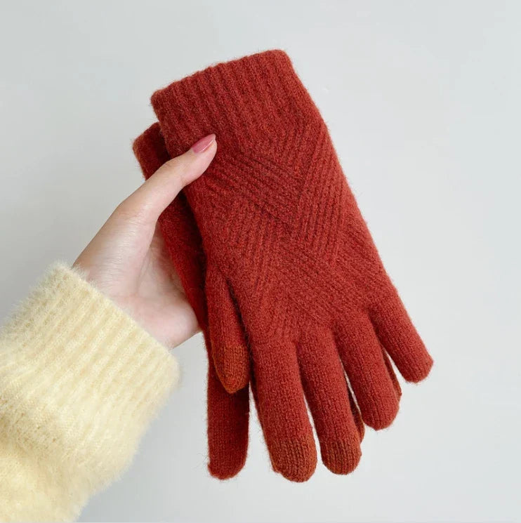 A hand in a fuzzy cream-colored sweater holds a pair of luxurious, rust-colored knit gloves made from a soft cashmere blend against a plain light background.