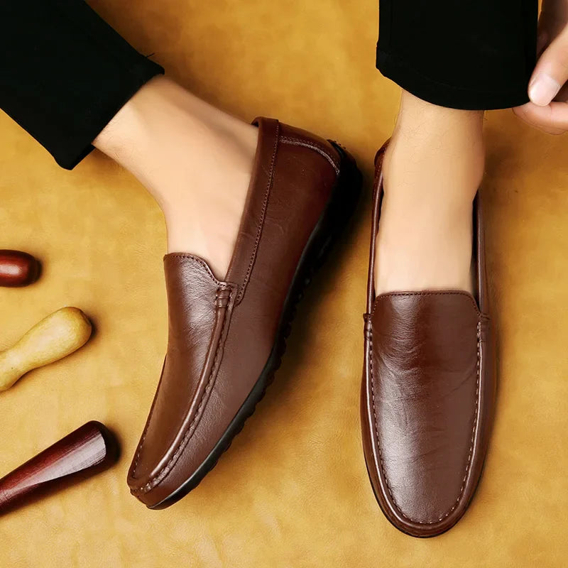 A pair of brown luxury shoes with a cushioned footbed worn by a person in black pants, shown from above on a tan background with wooden shoehorns nearby.