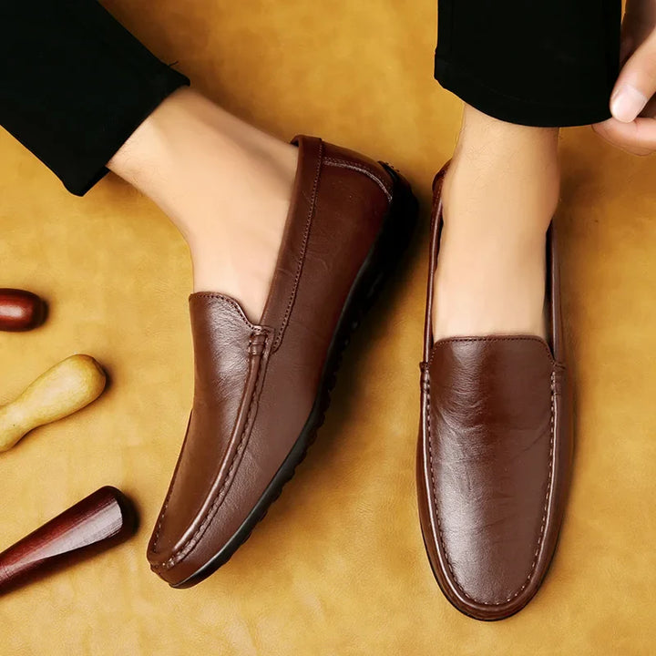 A pair of brown luxury shoes with a cushioned footbed worn by a person in black pants, shown from above on a tan background with wooden shoehorns nearby.