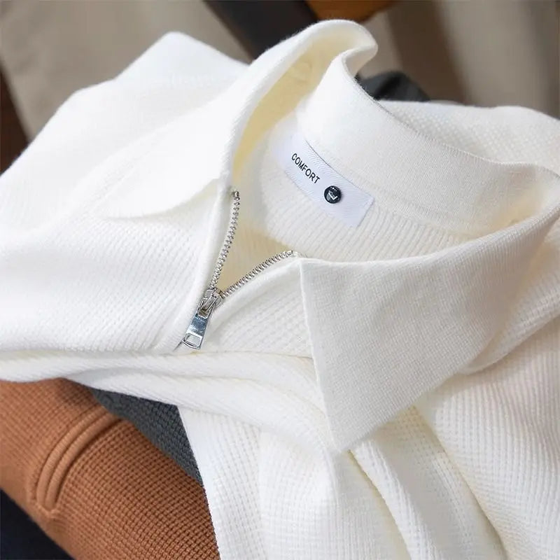 A close-up of a folded white ribbed zip-up collared sweater with a visible “CONSDDY” label, made from stretchy fabric, resting on top of brown and dark gray clothing.