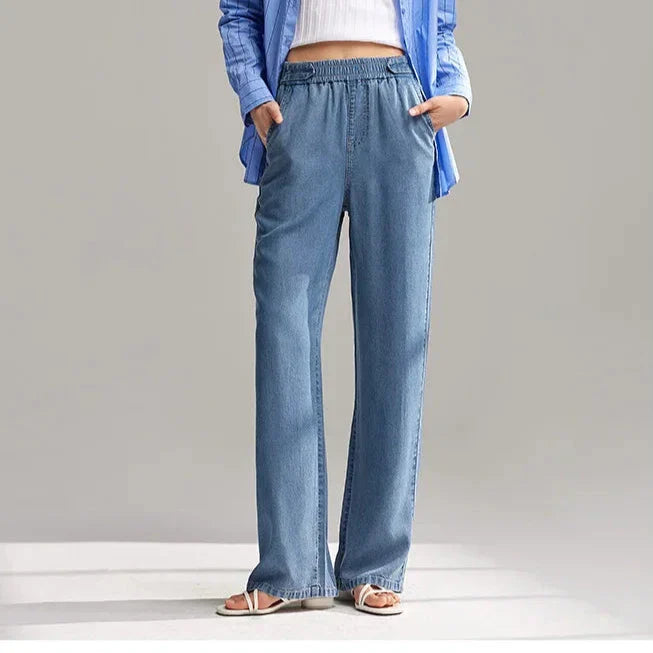 A person wearing wide-leg, light blue breathable denim pants with an elastic waistband, a white top, a blue shirt, and white sandals stands with hands in pockets against a plain background.