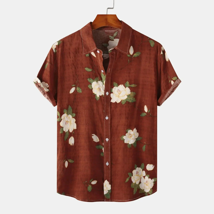 A short-sleeve, button-up linen shirt with a brown background featuring a white floral print and green leaves, displayed on a wooden hanger against a plain white background—perfect for effortless summer style.