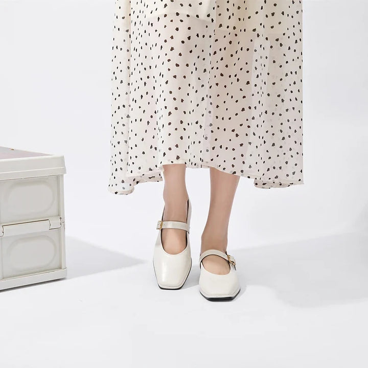 A person wearing a white, polka-dotted midi dress and sustainable shoes stands near a white box on a plain white background.