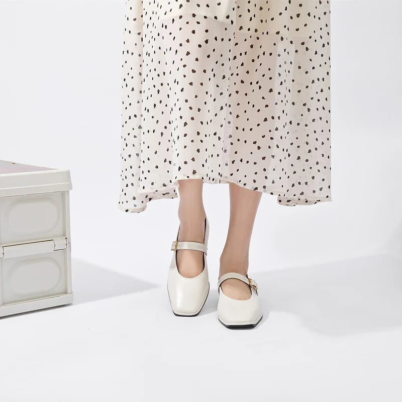 A person wearing a white, ankle-length dress with black polka dots and classic white Mary Jane shoes stands next to a white trunk against a plain background.