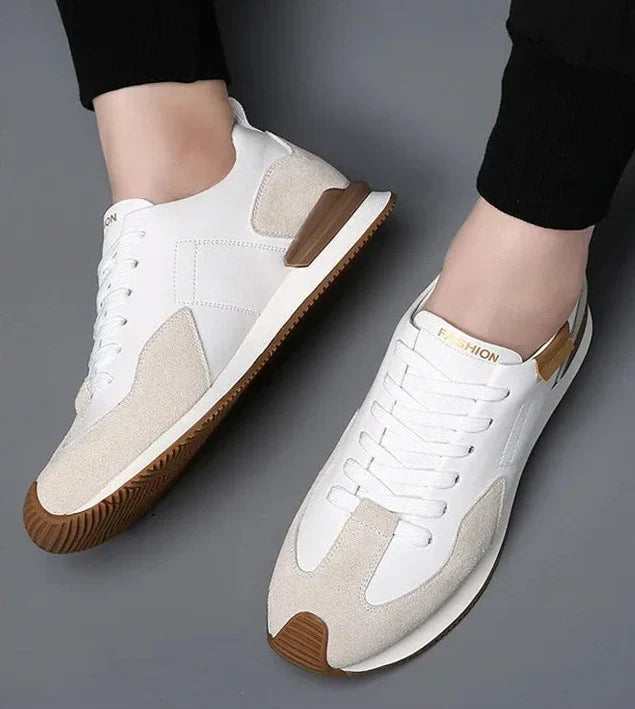 A person wearing white and beige sneakers with brown soles, labeled FASHION on the tongue, poses on a gray surface. These shoes feature top-grain leather and suede-like accents, offering both style and arch support.
