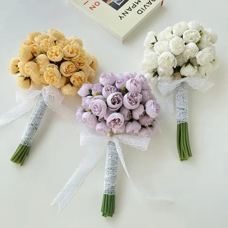 Three small bouquets of roses—yellow, light purple, and white—made with maintenance-free faux petals are wrapped in white lace and arranged on a white surface beside an open book.