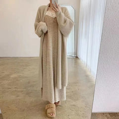 Person wearing a long, beige knit dress with a matching fluffy cardigan and fuzzy slippers, featuring a relaxed drape fit, standing indoors on a polished floor and posing in front of a mirror with sheer curtains in the background.