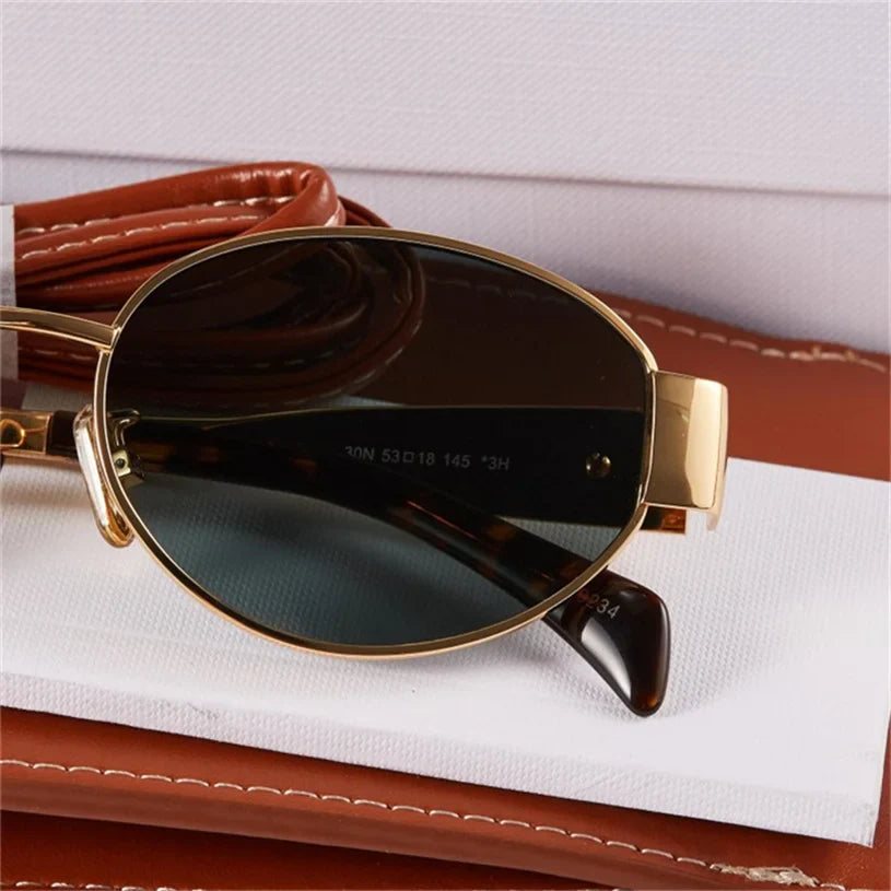 Close-up of vintage-inspired oval sunglasses with dark lenses and gold frames, resting on a folded brown leather case and a white cloth.