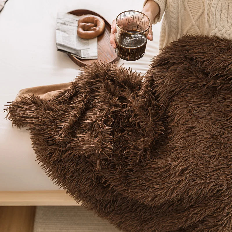 A person wearing a cream sweater sits on a bed covered with a soft faux fur throw blanket, holding a glass of dark beverage. A wooden tray with a newspaper and donut rests nearby, adding to the warm and cozy scene.