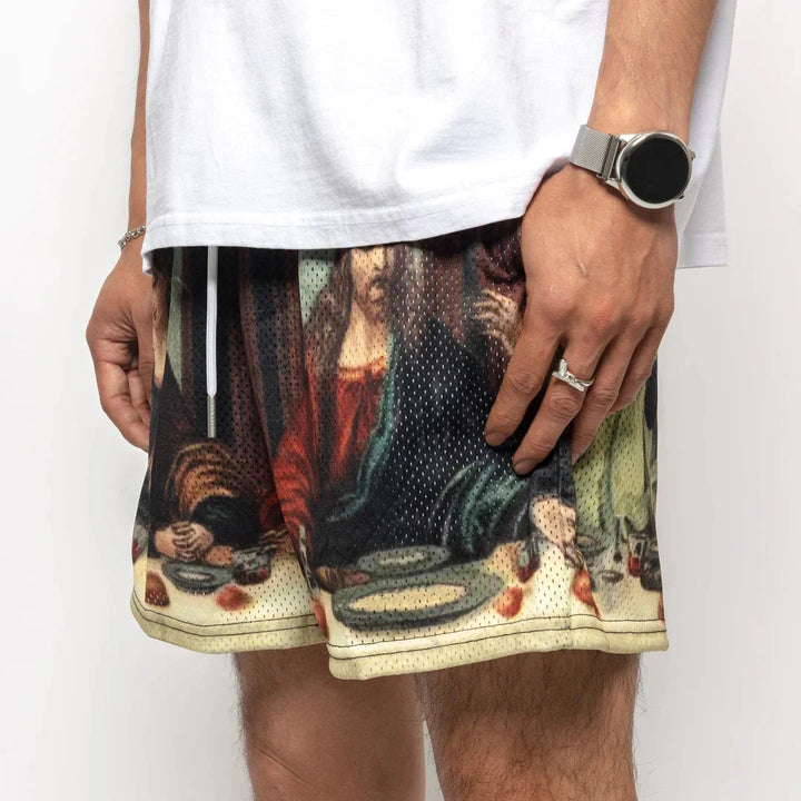 A person wears a white shirt and Last Supper graphic shorts featuring breathable mesh fabric with an elastic waistband. They also sport a wristwatch and ring, with their hand resting by their side.