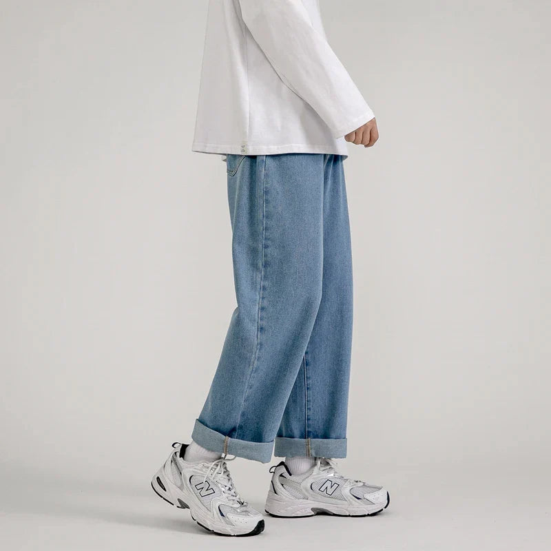 A person stands against a plain background wearing a white long-sleeve shirt, relaxed fit jeans in light blue with cuffs, white socks, and white New Balance sneakers. The persons head is not visible.