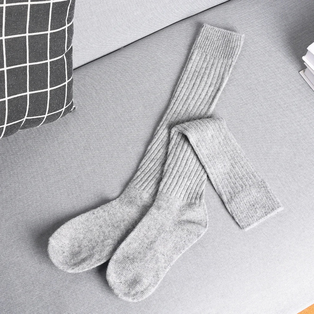 A pair of long, ribbed, gray cashmere cotton blend socks lies crossed on a light gray sofa next to a black and white checkered pillow and a stack of white papers.