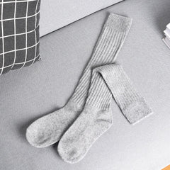 A pair of long, ribbed, gray cashmere cotton blend socks lies crossed on a light gray sofa next to a black and white checkered pillow and a stack of white papers.