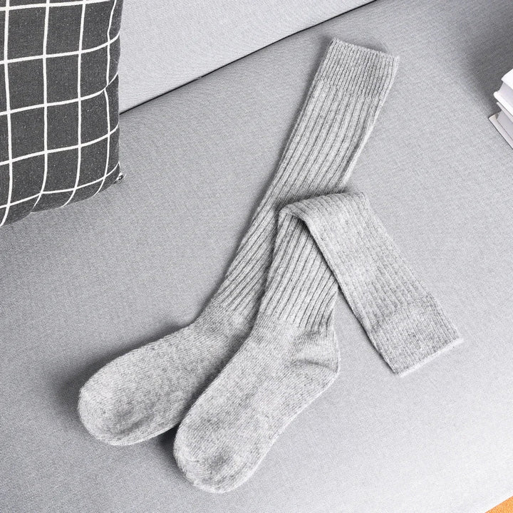A pair of long, ribbed, gray cashmere cotton blend socks lies crossed on a light gray sofa next to a black and white checkered pillow and a stack of white papers.
