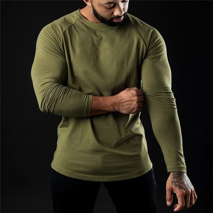 A man wearing a fitted, long sleeve stretch workout shirt in olive green and black pants stands against a black background, looking down and adjusting his right sleeve.