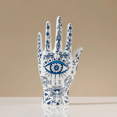 A white ceramic jewelry stand shaped like a hand, decorated with intricate blue patterns and an eye in the palm, serves as striking evil eye decor with floral motifs, standing upright on a smooth surface against a beige background.