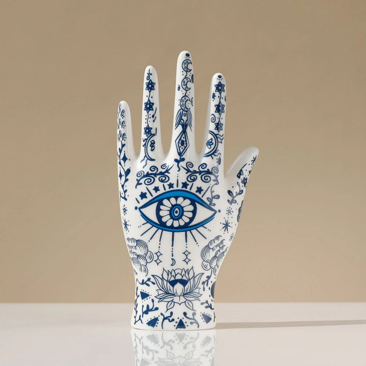 A white ceramic jewelry stand shaped like a hand, decorated with intricate blue patterns and an eye in the palm, serves as striking evil eye decor with floral motifs, standing upright on a smooth surface against a beige background.