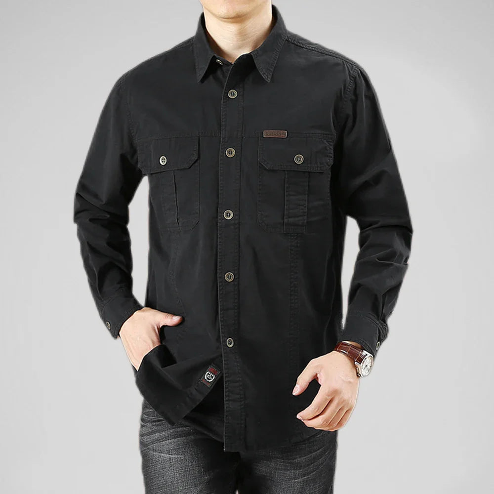 A person wearing a 100% cotton black button-up shirt with chest pockets and dark jeans stands with one hand in their pocket against a plain gray background.