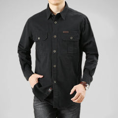 A person wearing a 100% cotton black button-up shirt with chest pockets and dark jeans stands with one hand in their pocket against a plain gray background.