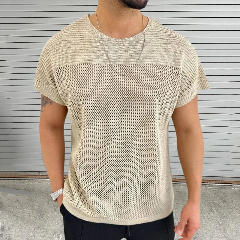 A man in a beige mesh knit tee with a breathable design, paired with a chain necklace and wristwatch, stands before a gray, horizontal-striped background. His face is not visible.