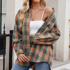 A woman wearing an oversized fit green and orange plaid button-down shirt over a white tank top and blue jeans stands outdoors near a building, with one side of the shirt off her shoulder.