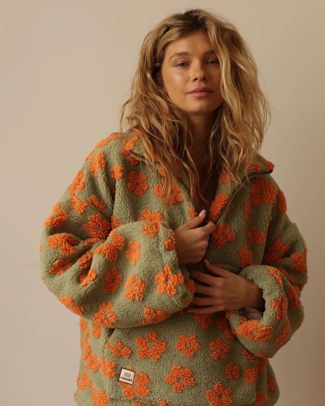 A woman with wavy blonde hair wears a green, extra-heavy fabric jacket patterned with large orange flowers, standing against a plain beige background. She looks at the camera with a soft expression, holding the jacket closed.