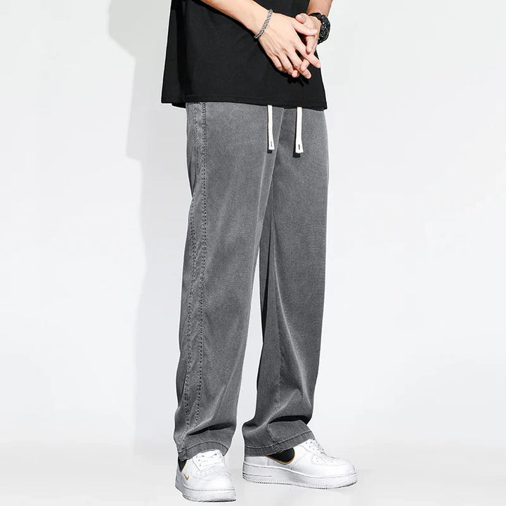 A person wearing gray Summer Loose Men Jeans jogger pants, a black shirt, a watch, and white sneakers stands against a plain white background. The photo captures them from the shoulders down.