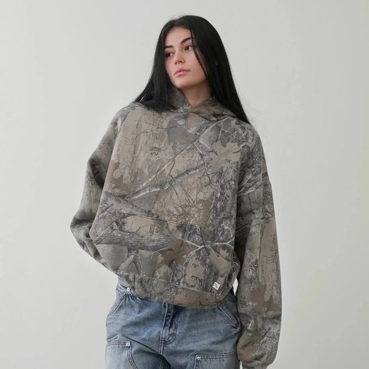 A person with long dark hair wears an oversized fit, camo pattern hoodie made from a cotton blend and blue jeans, standing against a plain light-colored background.