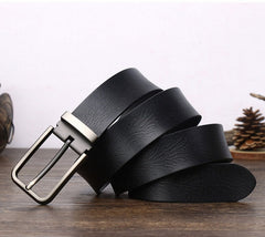 A black cowhide belt with a silver metal buckle is coiled on a wooden surface, with a pinecone and small decorative items blurred in the background.