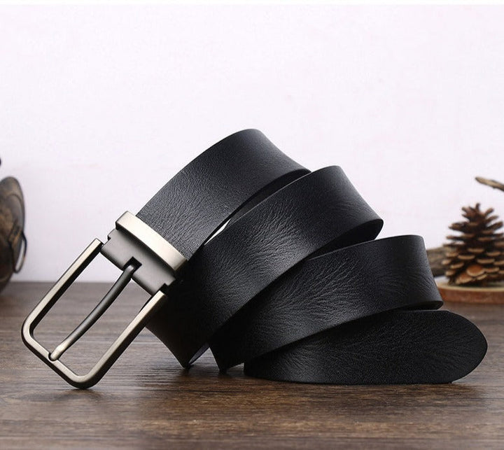 A black cowhide belt with a silver metal buckle is coiled on a wooden surface, with a pinecone and small decorative items blurred in the background.