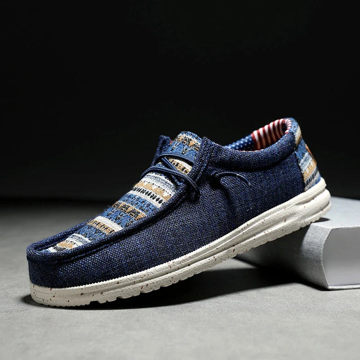 A single casual loafer-style shoe with a blue woven upper, featuring beige, white, and gray striped patterns, white speckled sole, and visible stitching, displayed on a gray surface against a dark background.