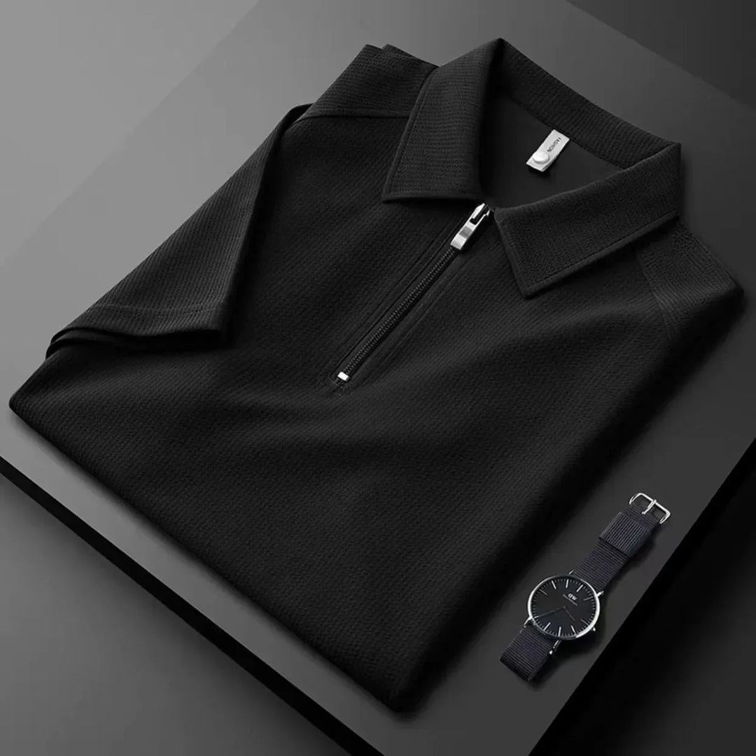A neatly folded black mens silk polo with a front zipper is placed on a black surface next to a minimalist black wristwatch with a silver bezel, highlighting the polos moisture-wicking comfort and modern style.
