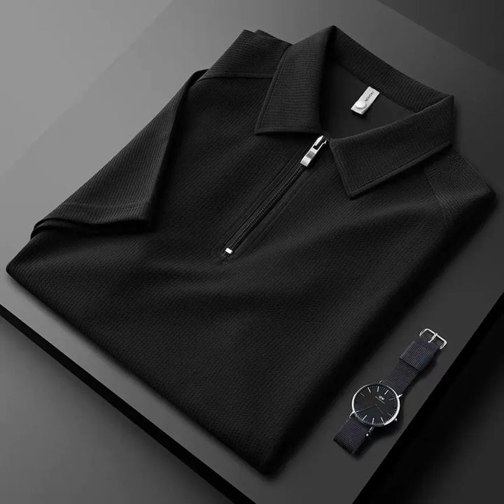 A neatly folded black mens silk polo with a front zipper is placed on a black surface next to a minimalist black wristwatch with a silver bezel, highlighting the polos moisture-wicking comfort and modern style.