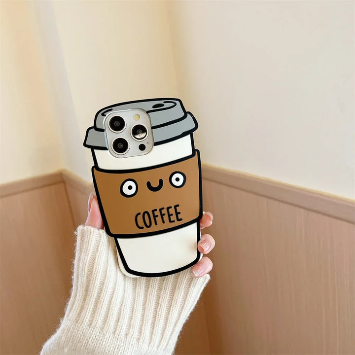 A hand in a white sweater holds a phone with a 3D coffee cup case made of soft silicone, featuring cartoon eyes, a smiling mouth, and the word COFFEE on a brown sleeve for shockproof protection.