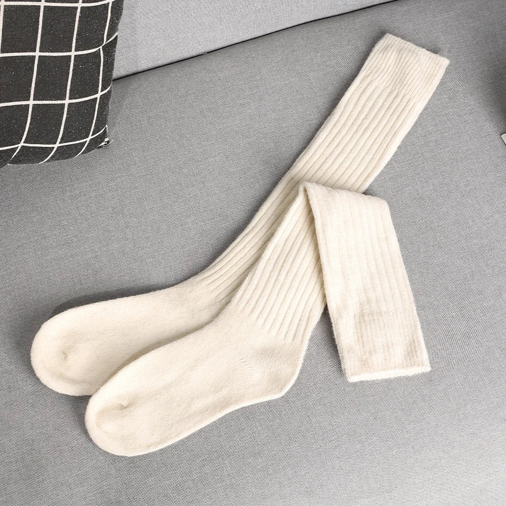 A pair of long, ribbed, cream-colored wool socks featuring superior insulation is laid out on a light grey couch beside a black and white checkered pillow.