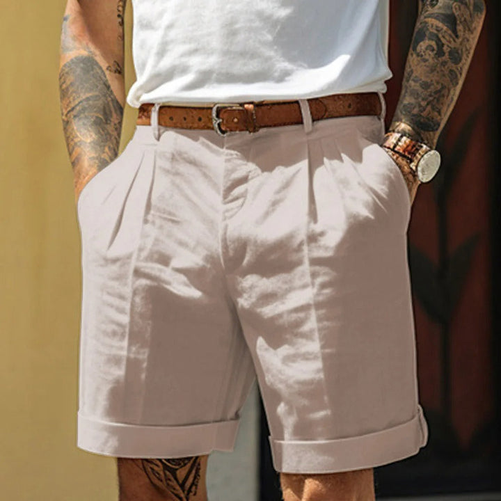 A person wearing light beige breathable linen shorts with cuffs, a brown leather belt, a white shirt tucked in, and a wristwatch. Their hands are in their pockets, and their arms have visible tattoos—perfect for a cool summer look.
