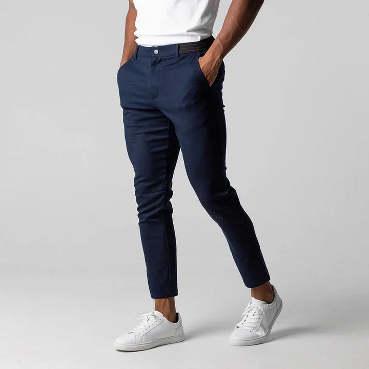 A person wearing a white t-shirt, navy blue slim fit ankle length pants with 4 way stretch, and white sneakers stands with hands in pockets against a plain light gray background.