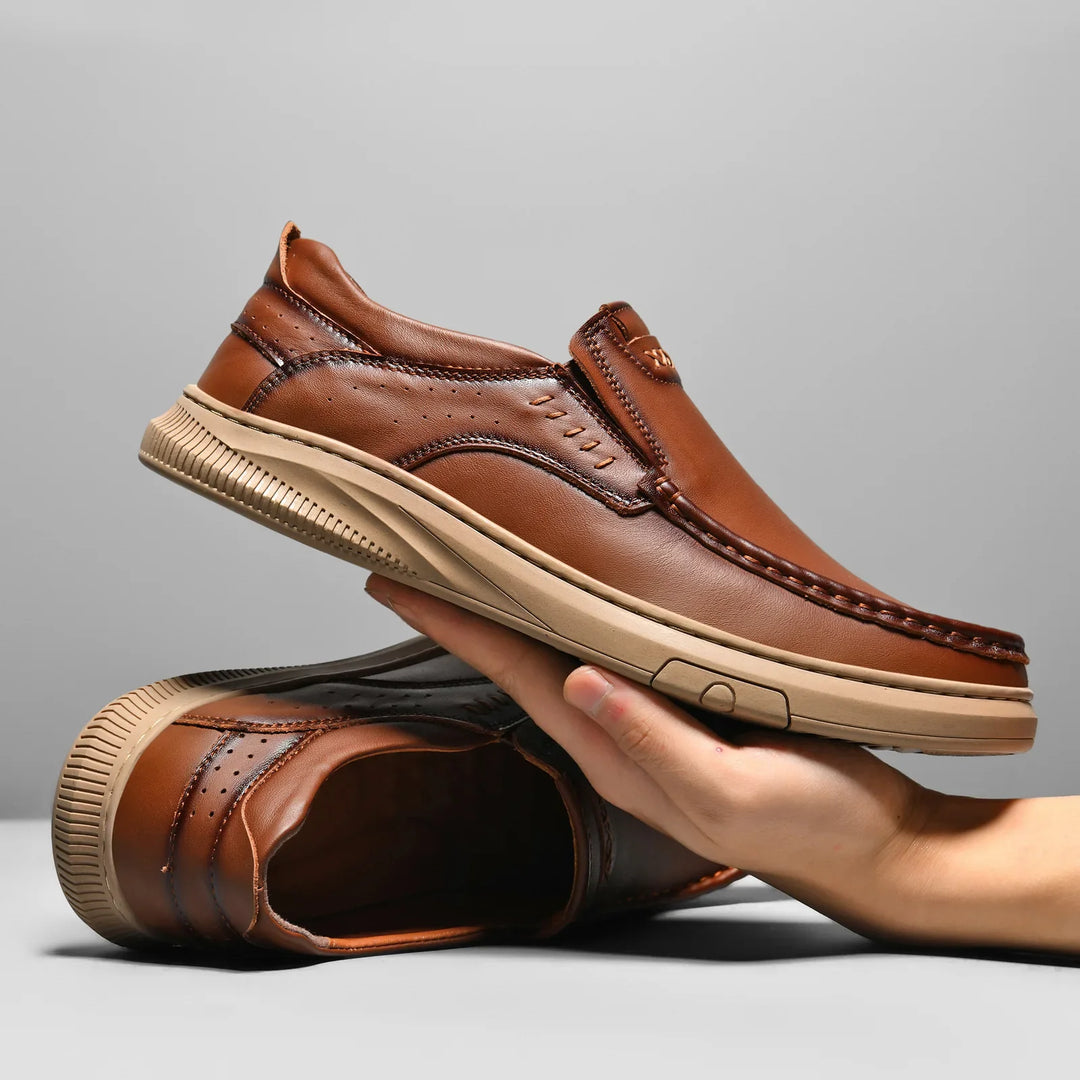 A hand holds a brown men's cowhide leather slip-on shoe with a tan soft sole. Another matching shoe lies on its side beneath, highlighting the stitched details and tread pattern against a plain gray background.